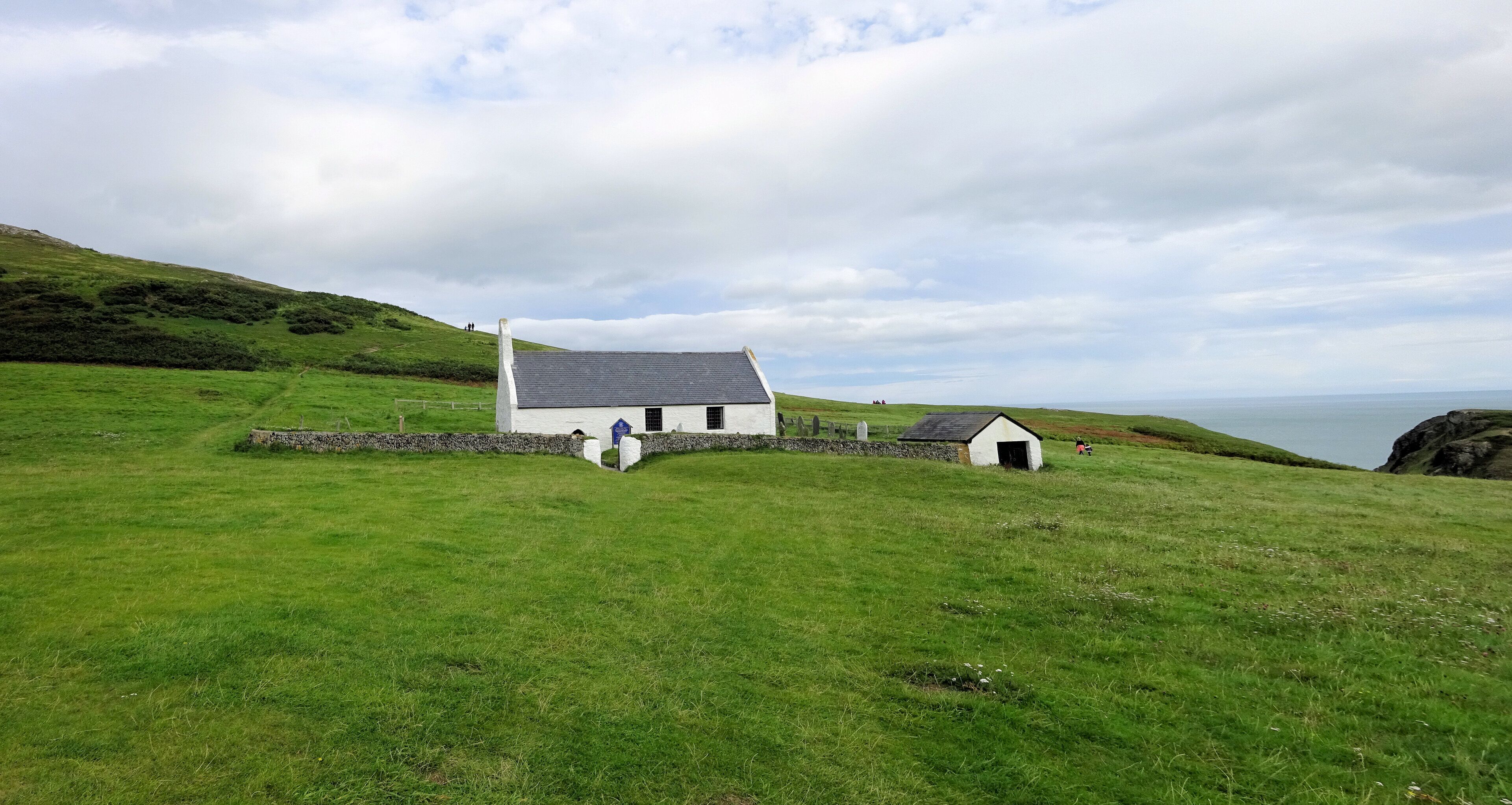 A white house with a grey roof on a grassy hill, a stone wall, and a small shed.