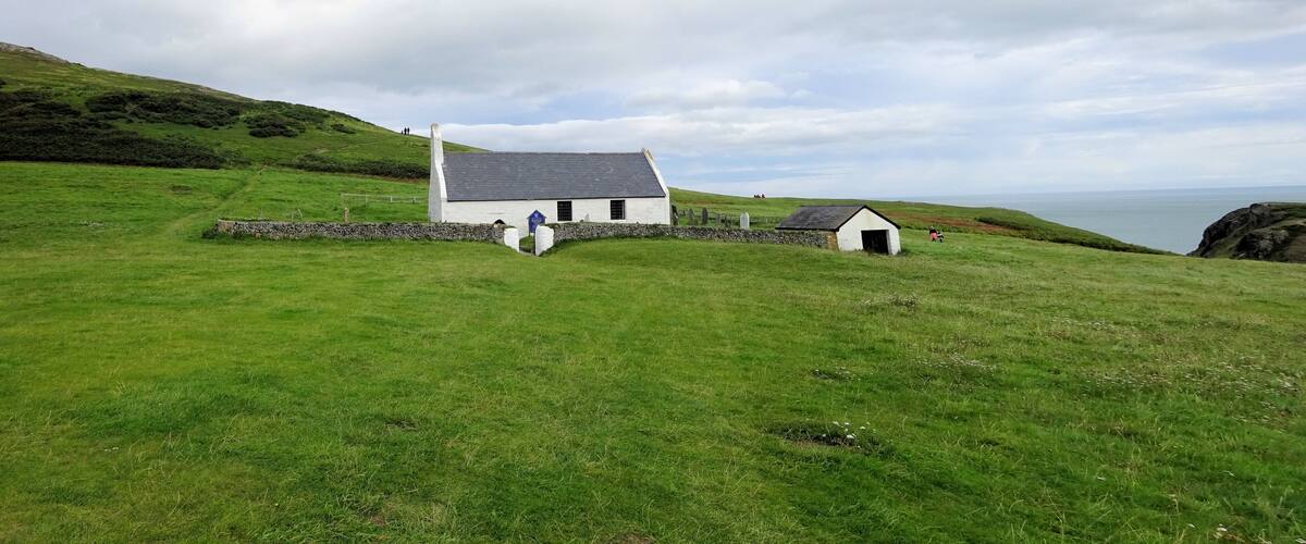A white house with a grey roof on a grassy hill, a stone wall, and a small shed.