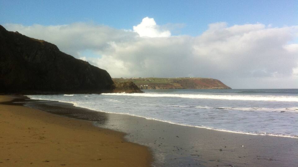 A beach with waves crashing onto the shore, a cliff, and a partly cloudy sky.