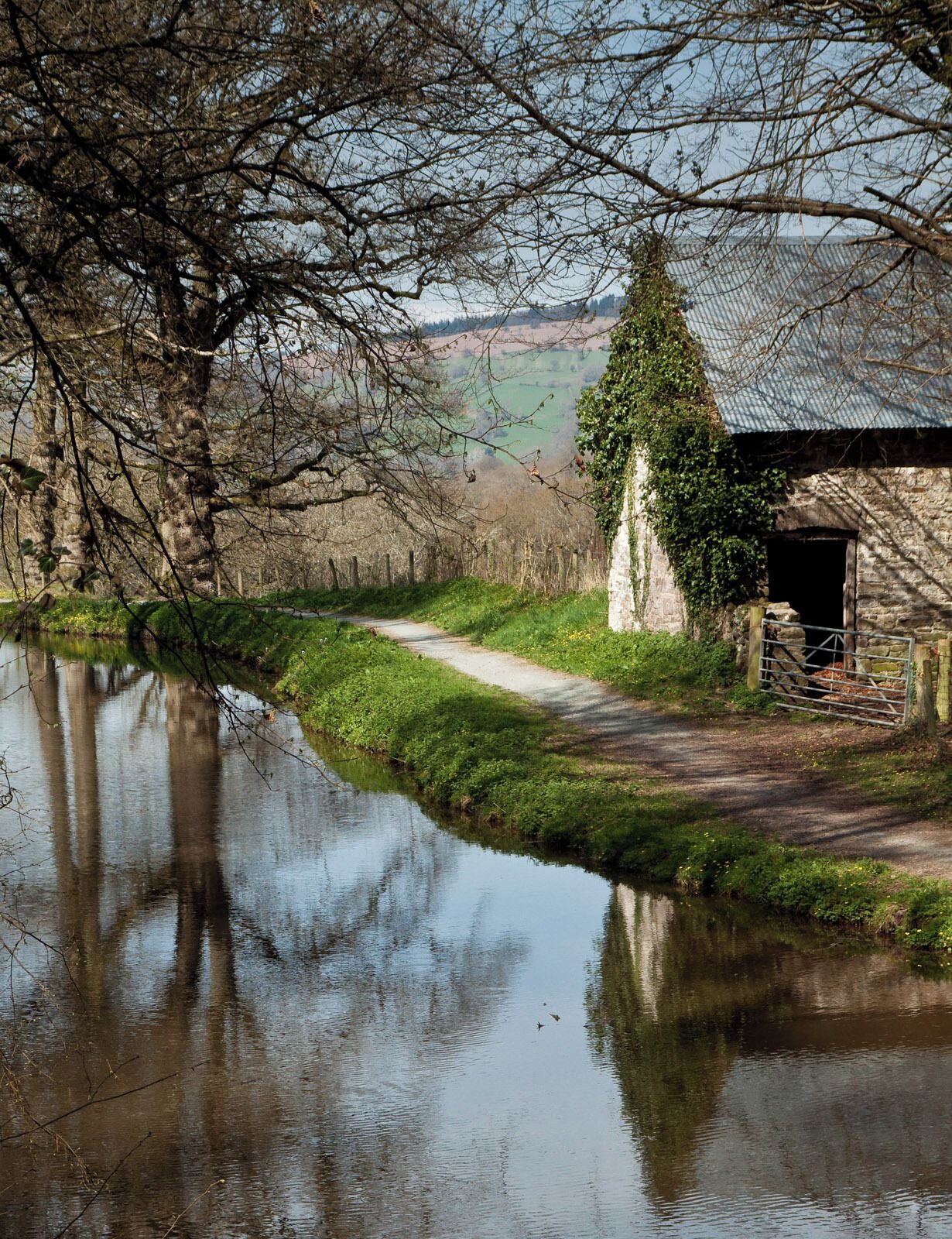A rural landscape with a narrow path beside a calm river, a stone building with a metal gate, and a tree with ivy.