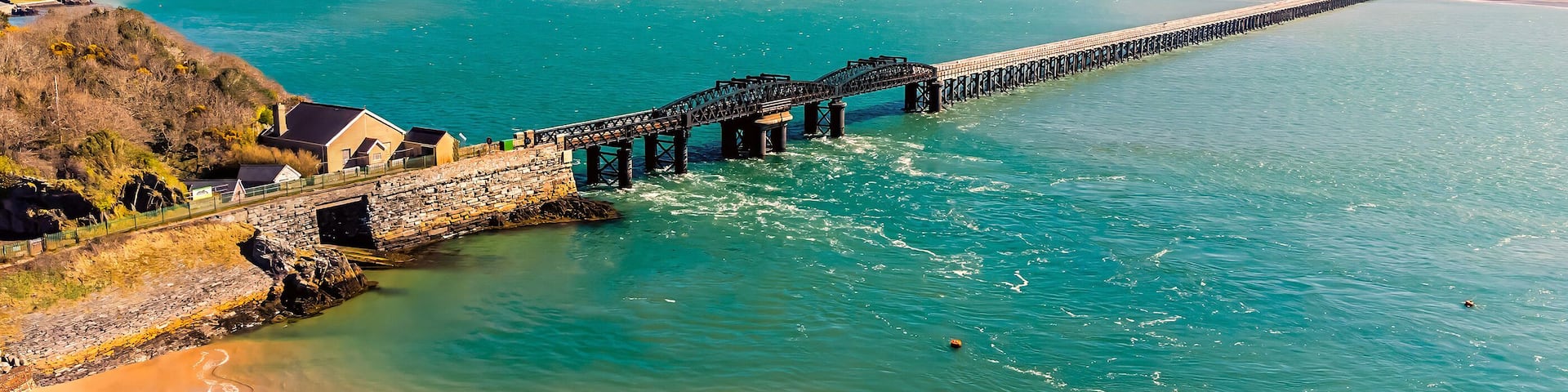A coastal landscape with a long pier extending into turquoise waters, surrounded by hills and a sandy beach.
