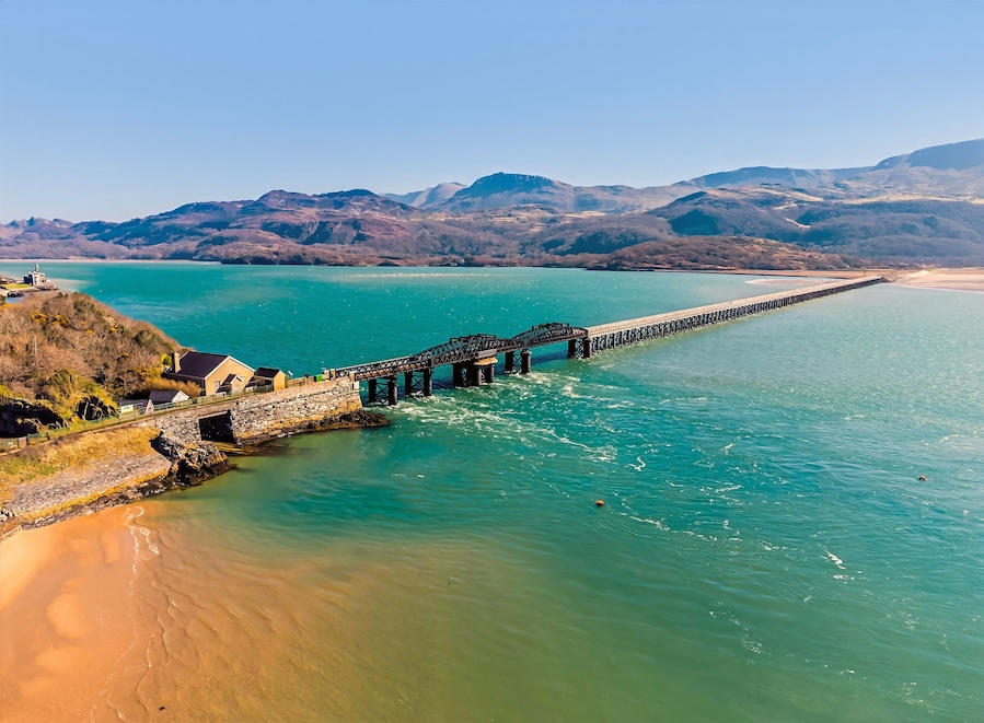 A coastal landscape with a long pier extending into turquoise waters, surrounded by hills and a sandy beach.