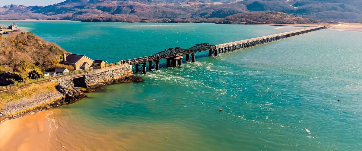 A coastal landscape with a long pier extending into turquoise waters, surrounded by hills and a sandy beach.