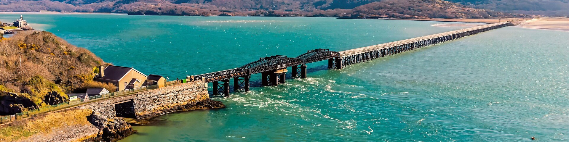 A coastal landscape with a long pier extending into turquoise waters, surrounded by hills and a sandy beach.