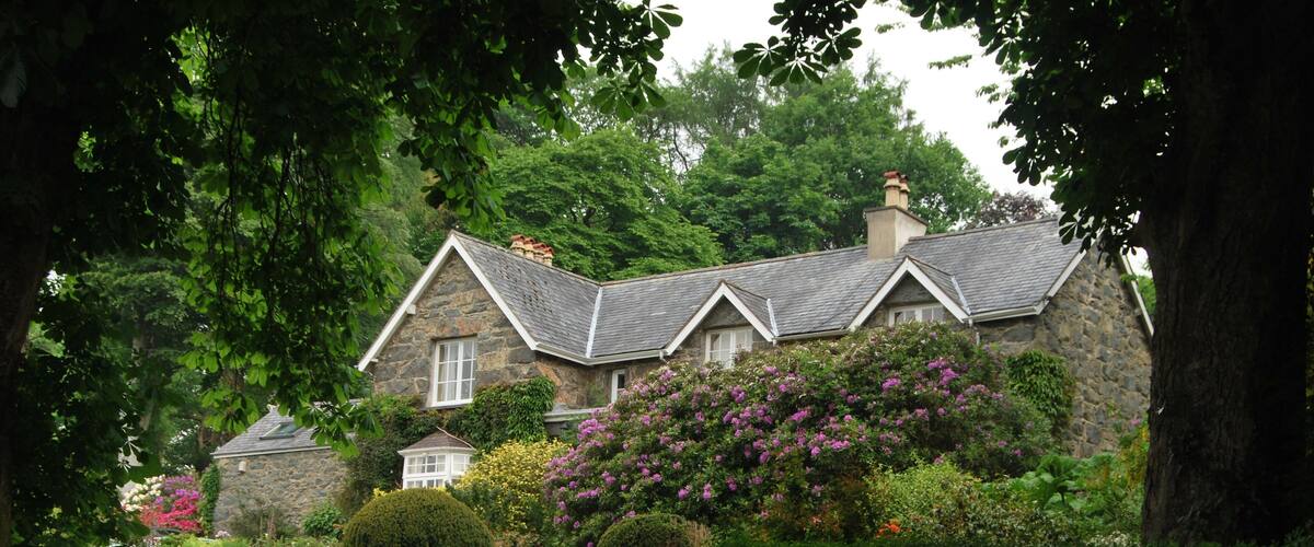 A stone house with a sloped roof, surrounded by lush greenery and flowering plants.