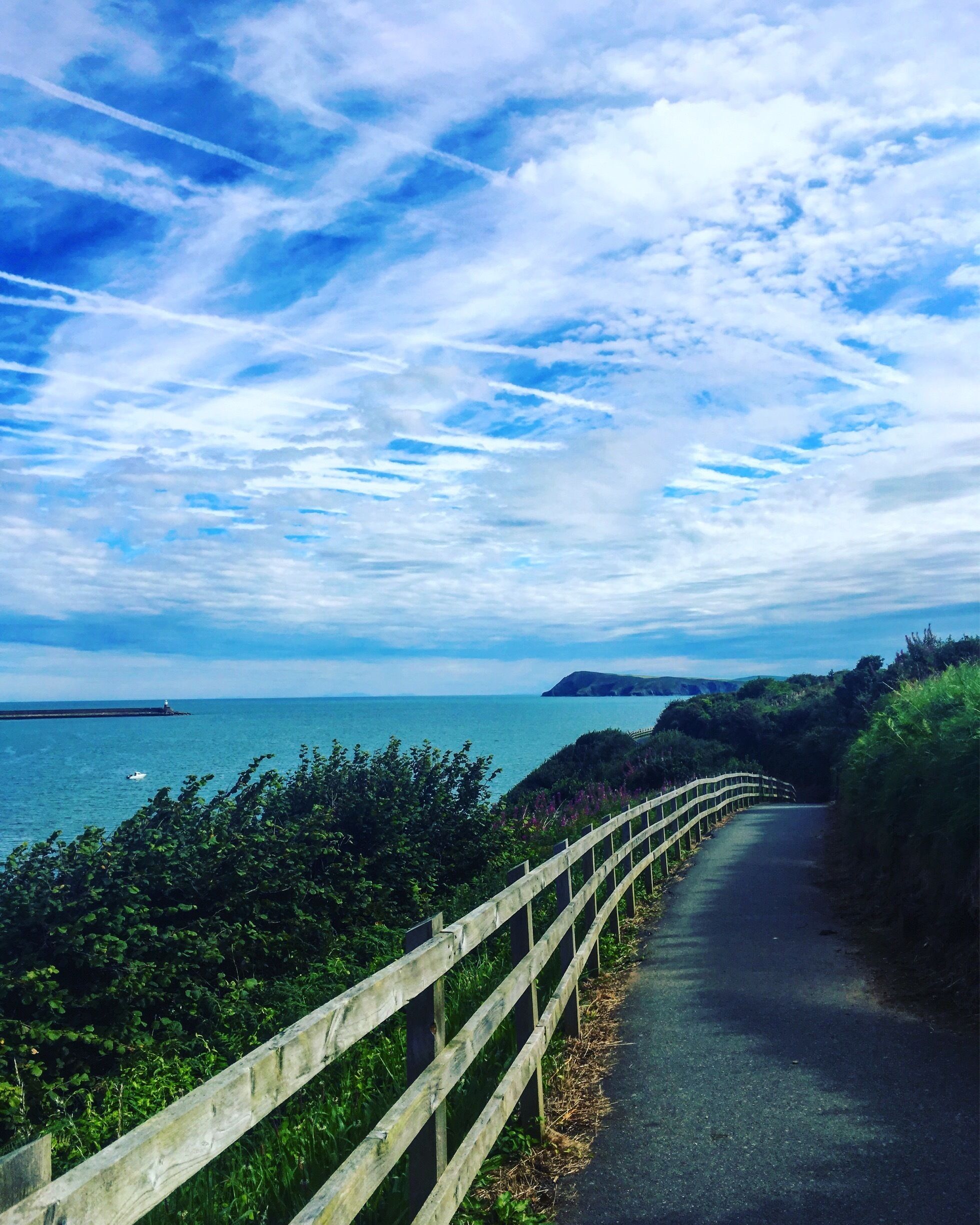 A coastal road with a wooden fence, a lighthouse, and a view of the sea and distant hills.