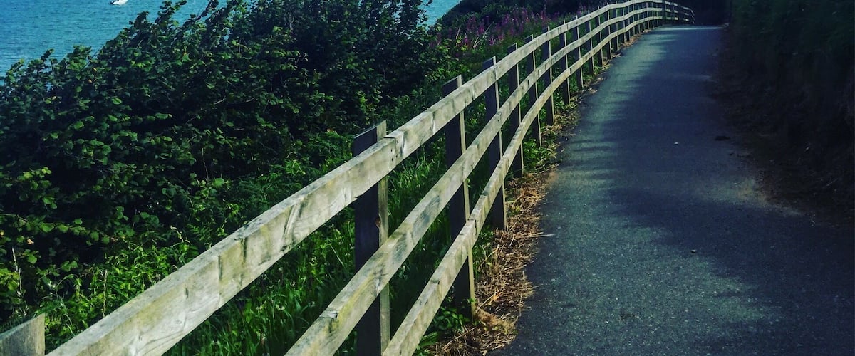A coastal road with a wooden fence, a lighthouse, and a view of the sea and distant hills.