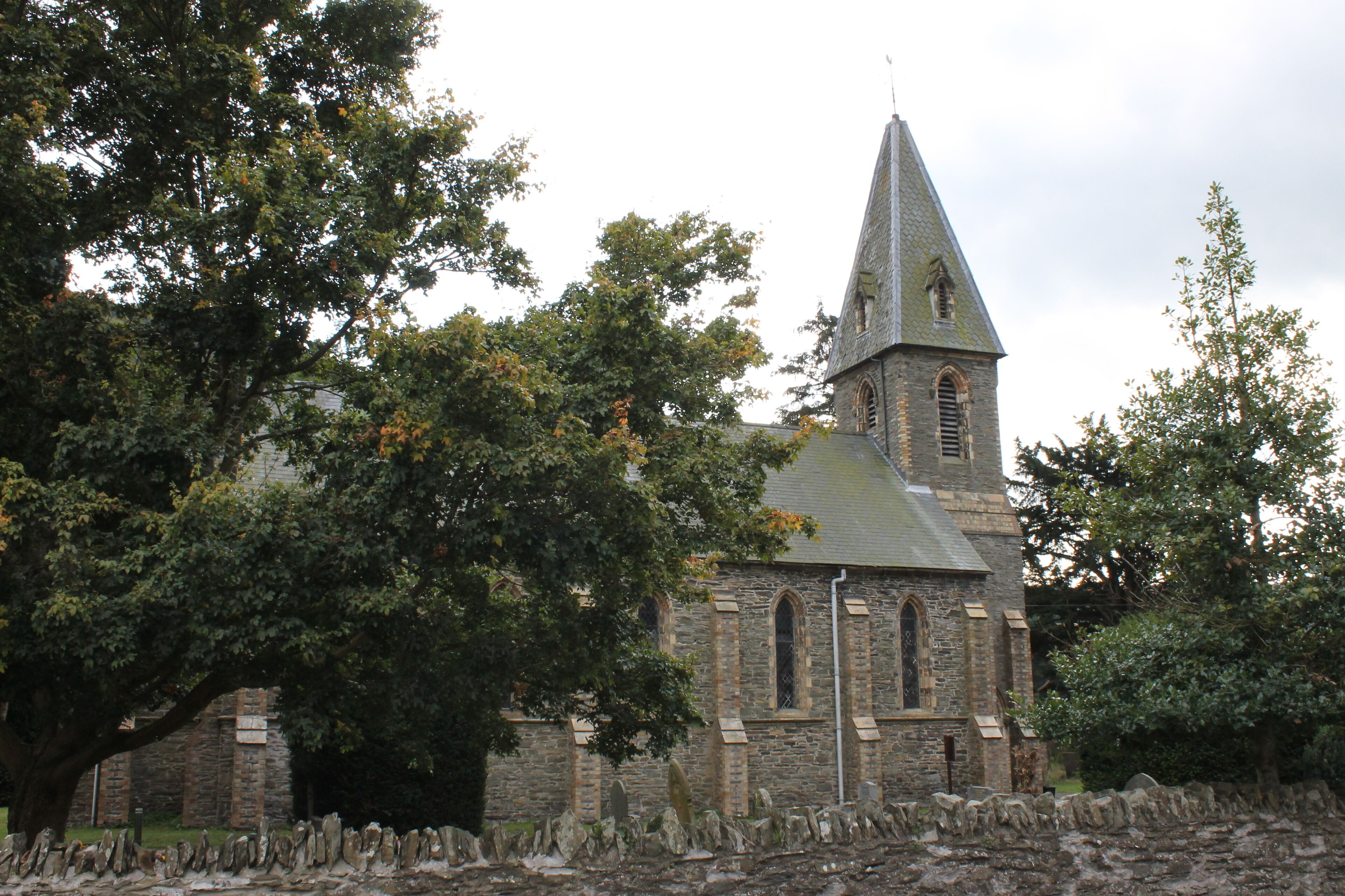 A stone church with a pointed steeple and arched windows, surrounded by trees and a stone wall.