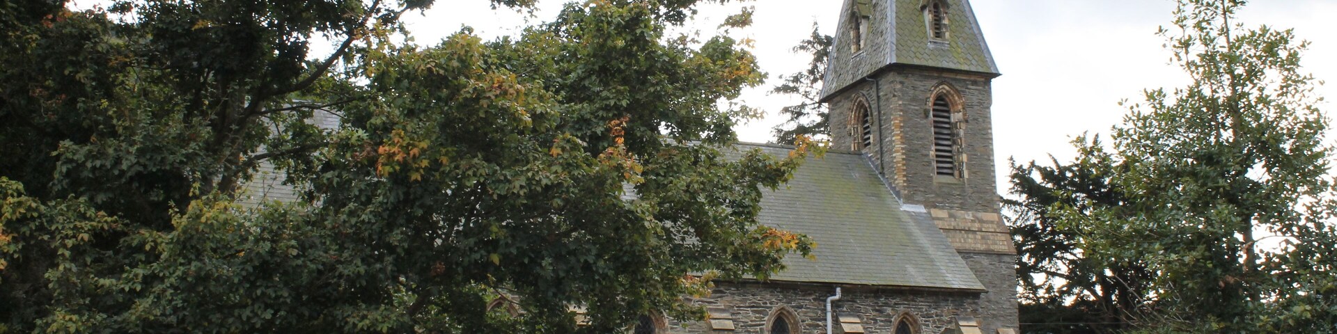 A stone church with a pointed steeple and arched windows, surrounded by trees and a stone wall.
