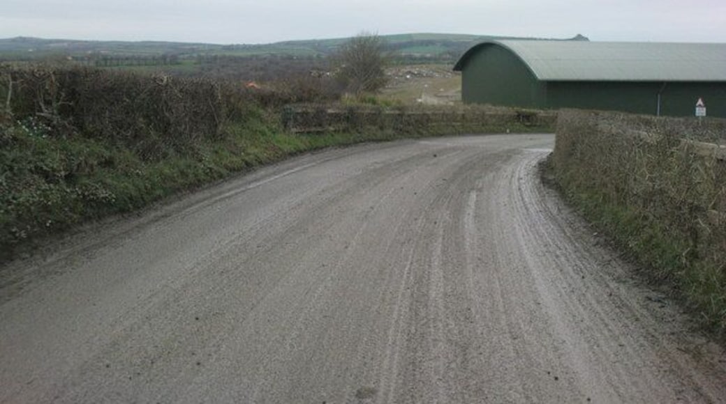 Un camino rural se curva hacia la derecha, bordeado por muros de piedra y vegetación, conduciendo a un edificio de techo verde.