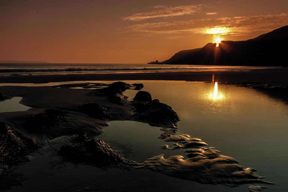 A sunset over a rocky beach with calm water reflecting the sky.