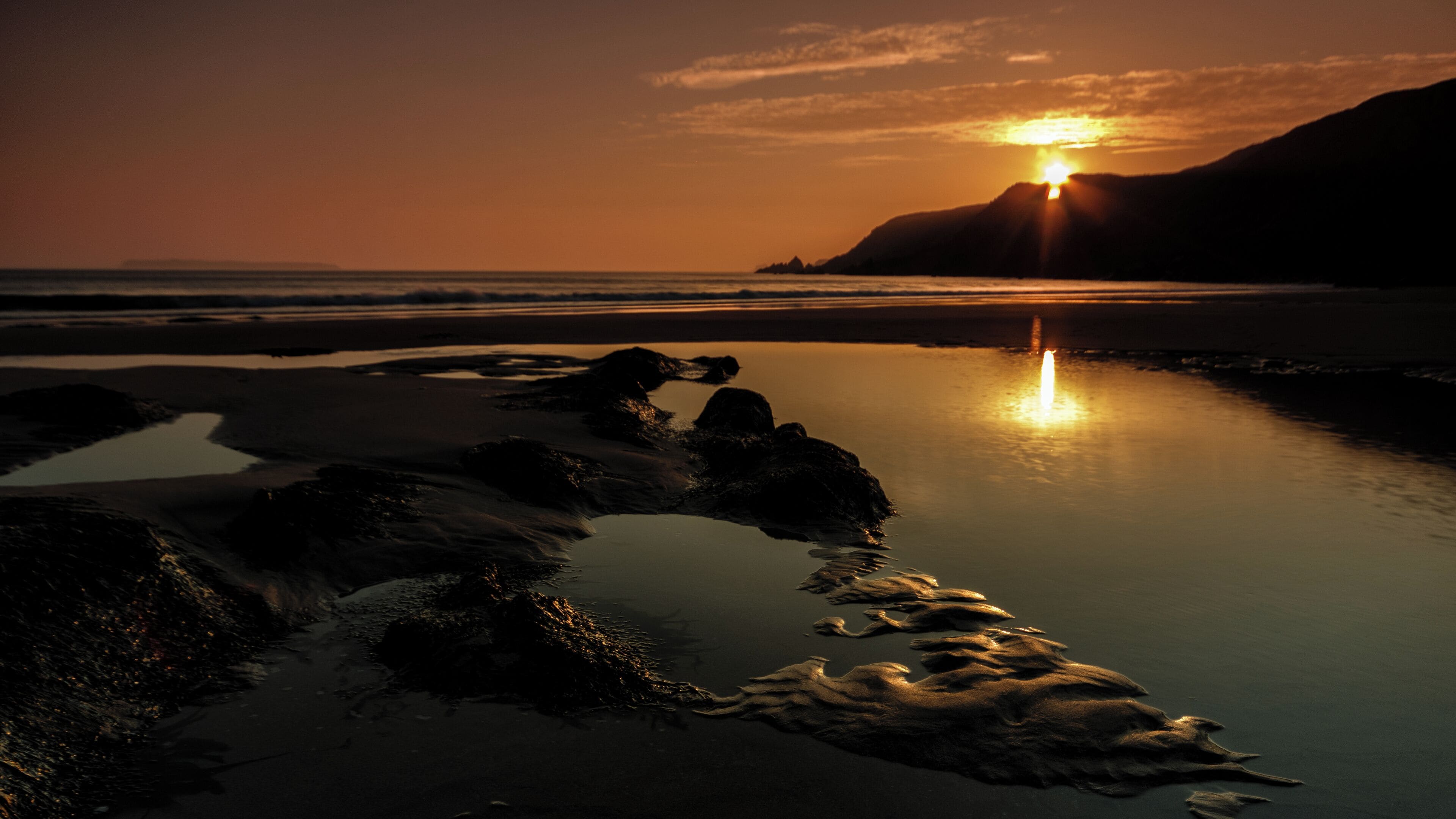 A sunset over a rocky beach with calm water reflecting the sky.