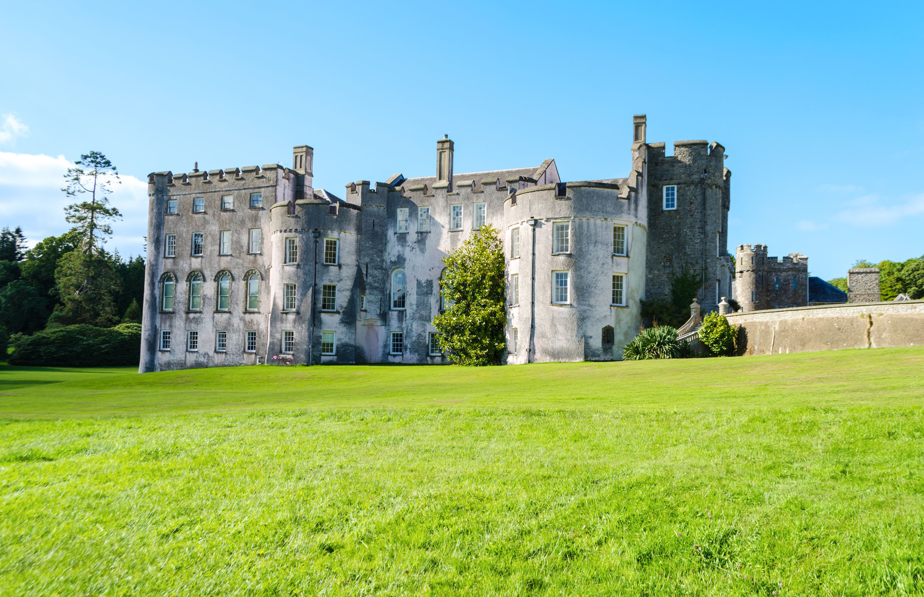 A large, historic castle with multiple towers and turrets, surrounded by a well-maintained lawn and trees.