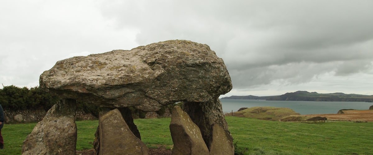 A large standing stone structure in a grassy field with a body of water and hills in the background.