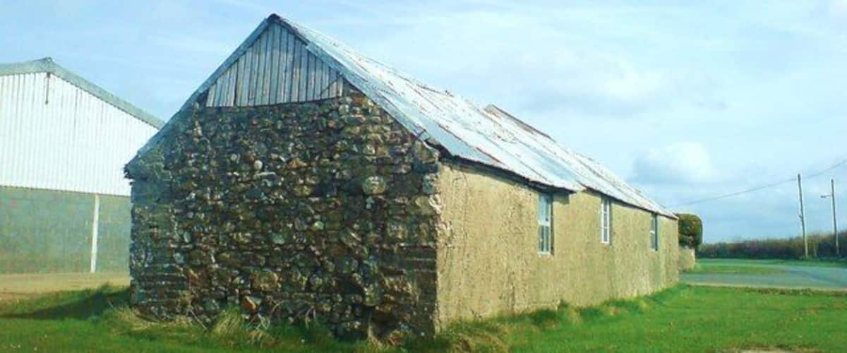A stone barn with a metal roof, surrounded by a grassy field and a white building in the background.