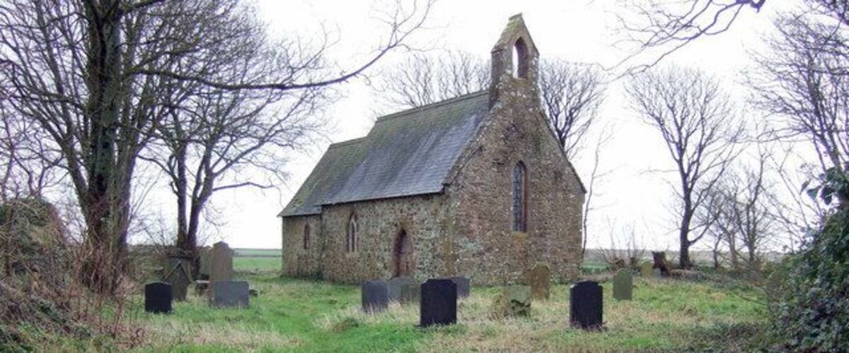 A stone church with a steep roof and a bell tower, surrounded by a grassy field and several headstones.