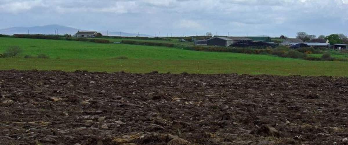 A rural landscape with a plowed field, green fields, and distant buildings under a cloudy sky.