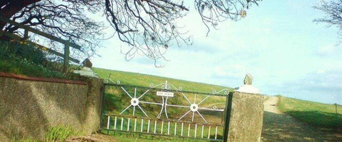 A gated entrance with a metal gate, a stone wall, and a grassy hill in the background.