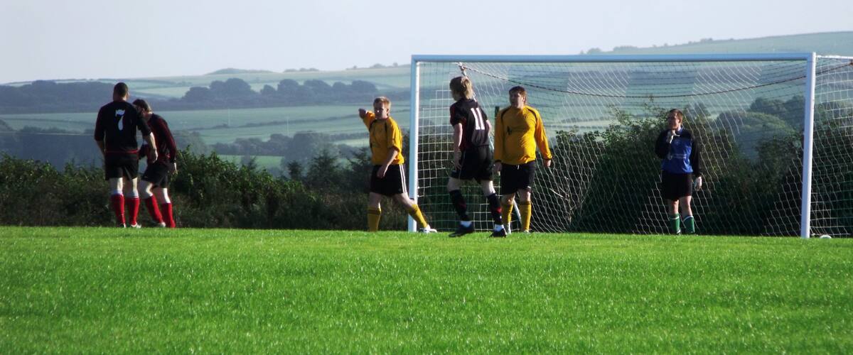 A soccer field with players in black and yellow jerseys, a goalpost, and a rural landscape in the background.