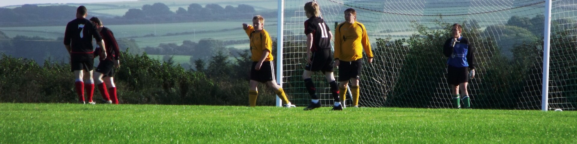 A soccer field with players in black and yellow jerseys, a goalpost, and a rural landscape in the background.
