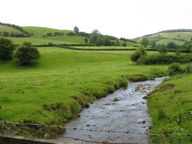 A rural landscape with a stream, green fields, and rolling hills.