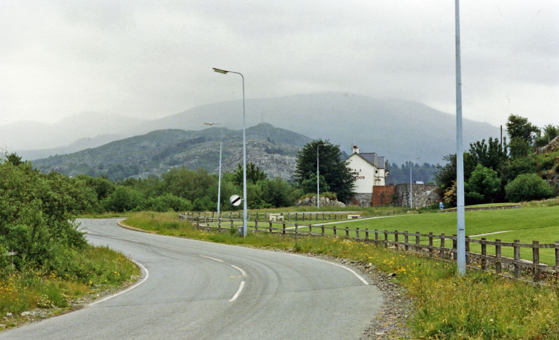 Near site of Cwm-y-glo station towards Llanberis Pass. View SE on A4086 near Llyn Padarn, the railway having been off frame to the left: ex-LNW Caernarvon - Llanberis branch. The station was closed to regular passenger services from 22/9/30 (goods 29/6/64), but the excursions ran to Llanberis until 7/9/62 (goods 7/12/64). This is the way to Snowdon and its Mountain Railway - off to the right: in the centre background is Elidir Fawr (3,026 ft.).
