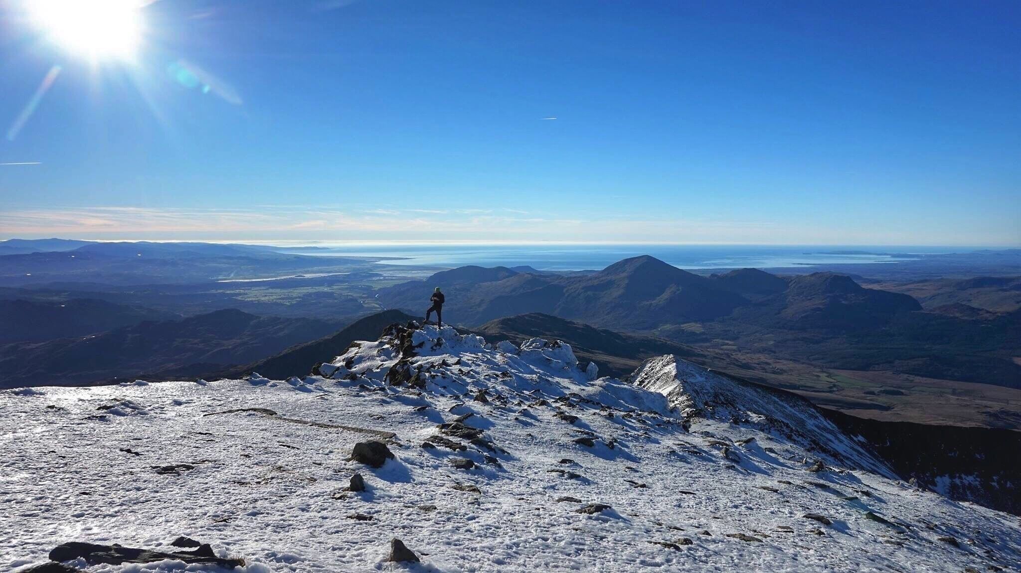 A snowy mountain peak with a person standing on it, overlooking a vast landscape with distant mountains and a clear sky.