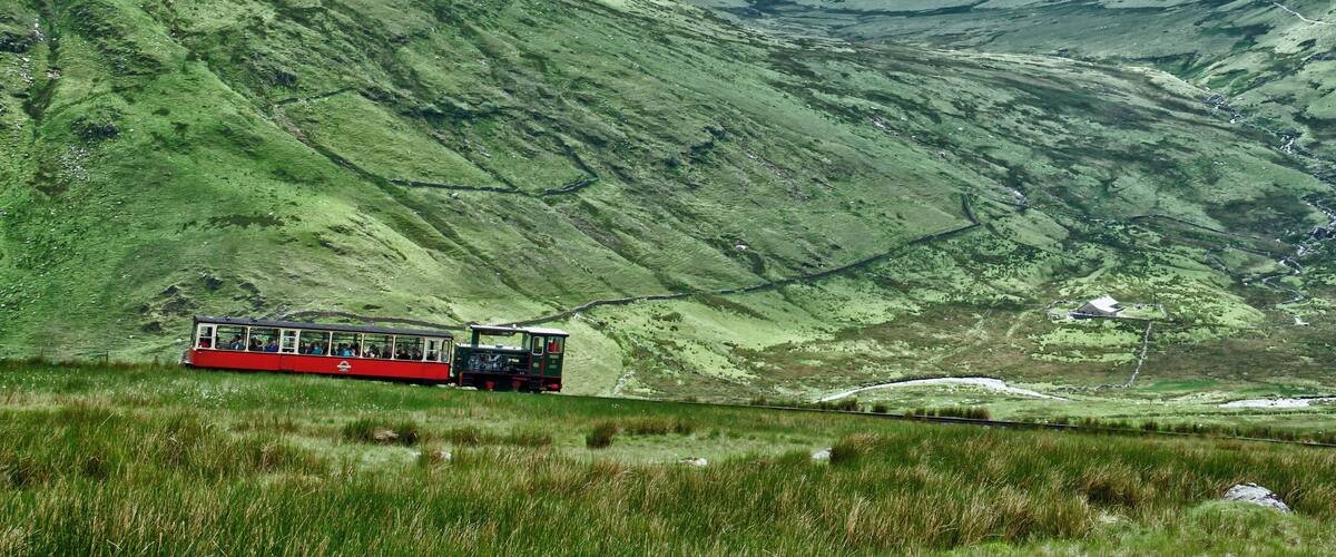 A red train on a mountain track with lush green hills.