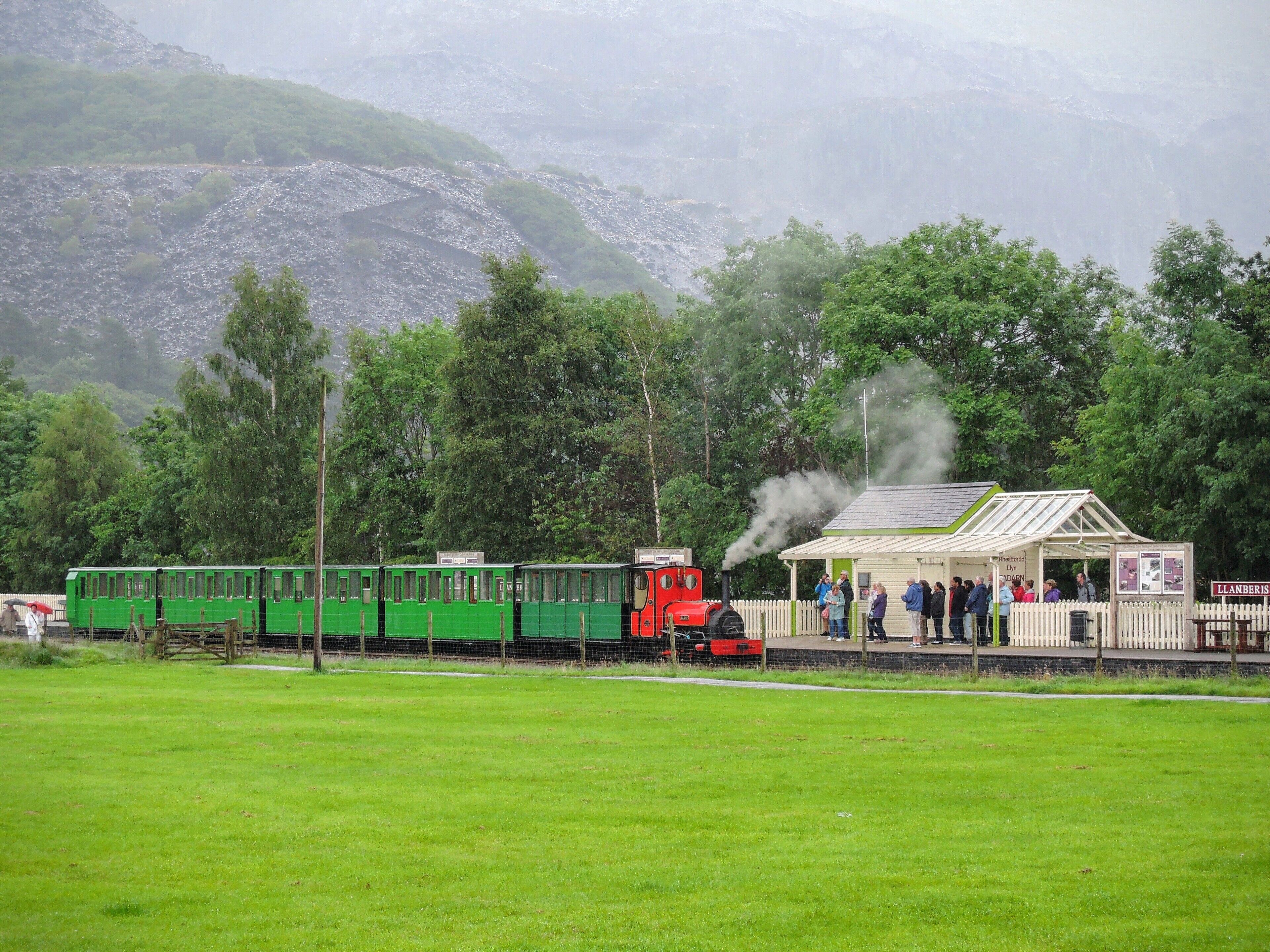 A green train with a red locomotive at a station surrounded by lush greenery and mountains.