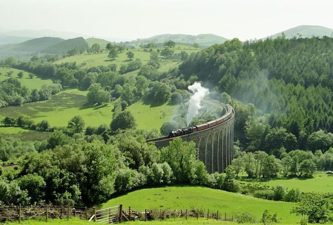 A steam train on a bridge through a green landscape.