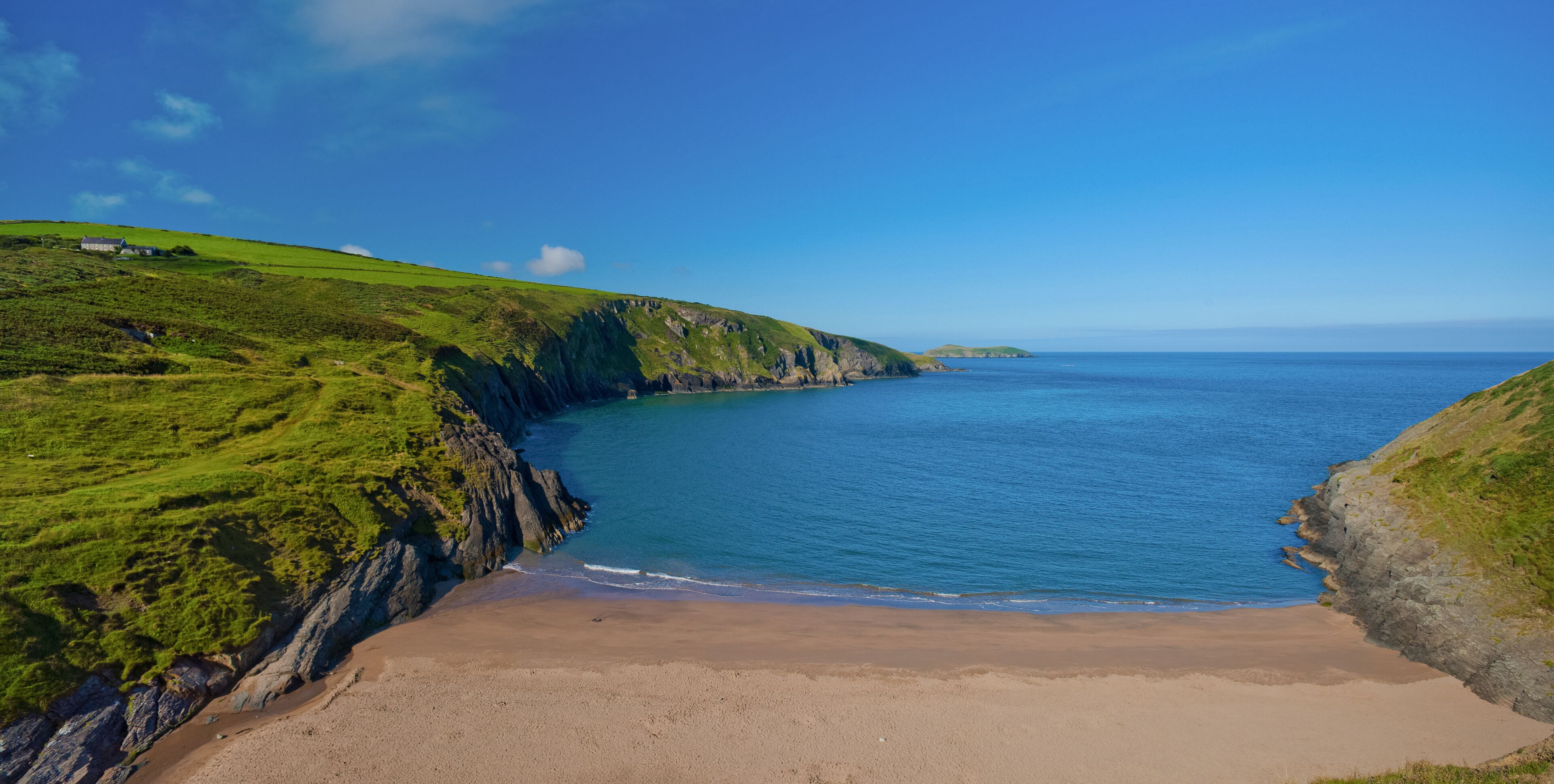 A coastal landscape with a sandy beach, green hills, and a clear blue sea.