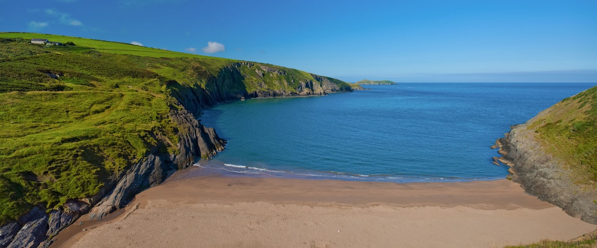A coastal landscape with a sandy beach, green hills, and a clear blue sea.
