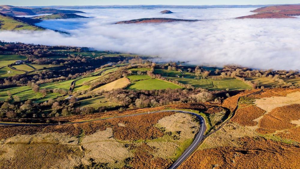 A winding road through rolling hills with fields and a blanket of clouds below.