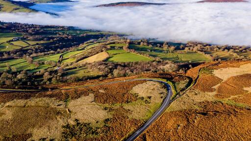 A winding road through rolling hills with fields and a blanket of clouds below.