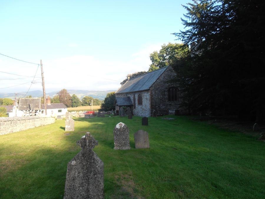 A rural churchyard with gravestones, a stone building, and a red vehicle in the background.