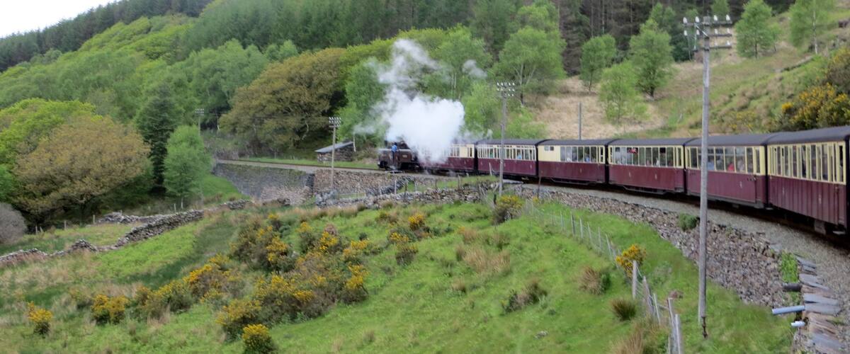 A steam train with passengers traveling through a rural landscape with green hills and trees.