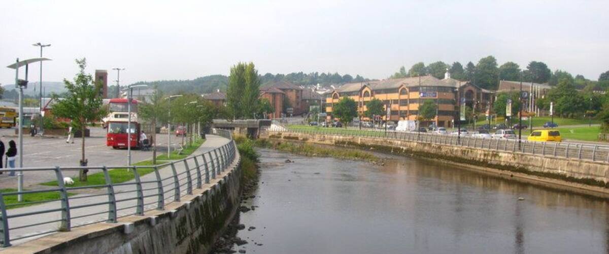 A river with a concrete embankment, a red bus, and buildings in the background.