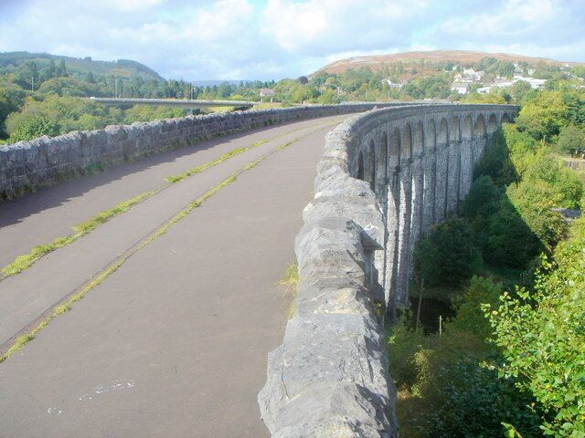A stone viaduct with multiple arches over a river, surrounded by greenery and hills in the background.