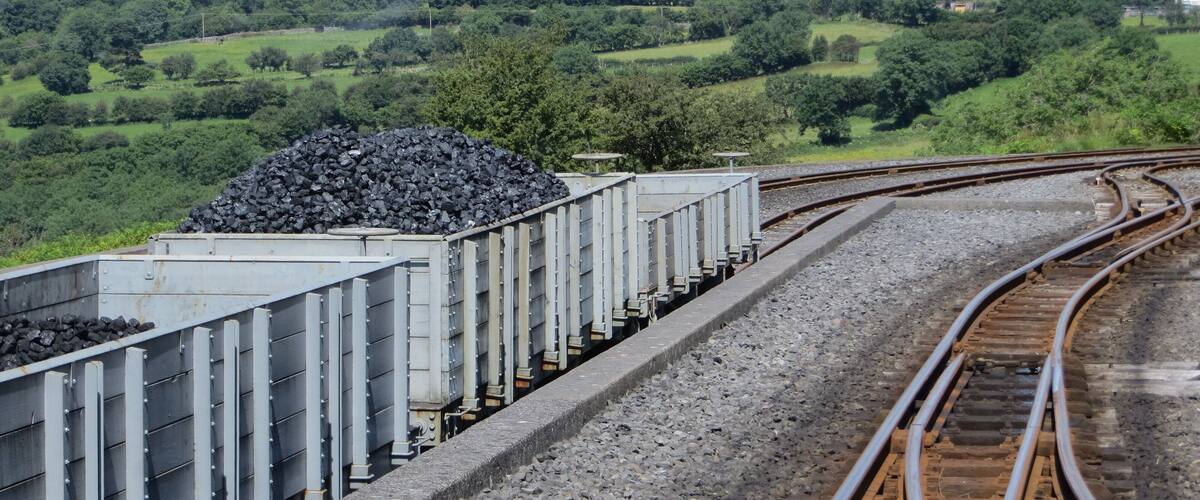 A train carrying coal cars on tracks with a green landscape in the background.