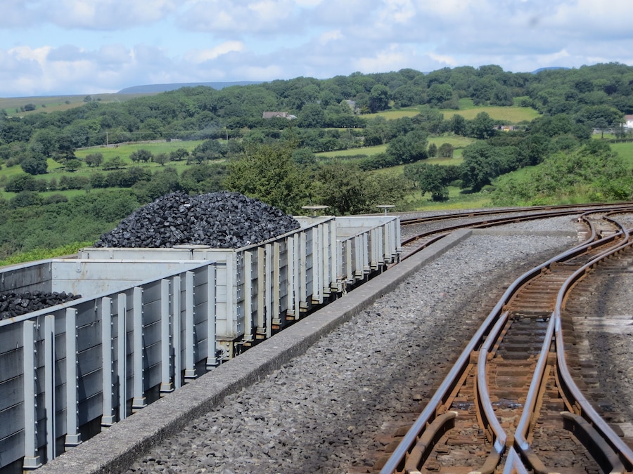 Um trem transportando vagões de carvão sobre trilhos, com uma paisagem verde ao fundo.
