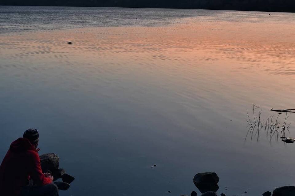 Una persona sentada sobre una roca junto a un lago tranquilo al atardecer.