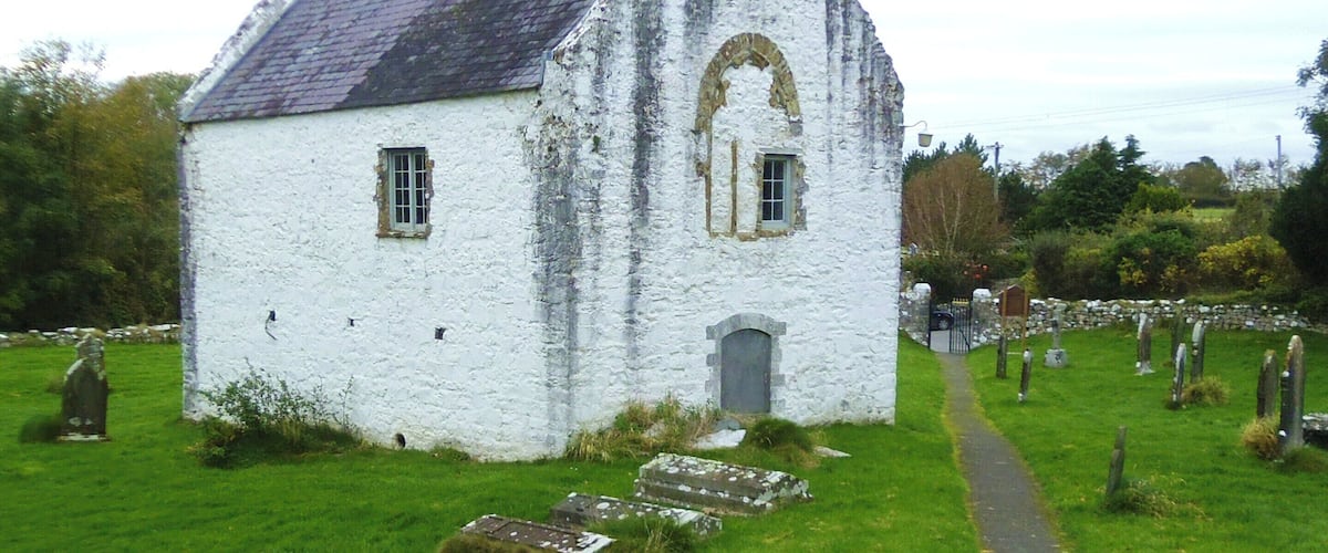 A white stone house with a steep roof and arched windows, surrounded by a grassy field and a cemetery.