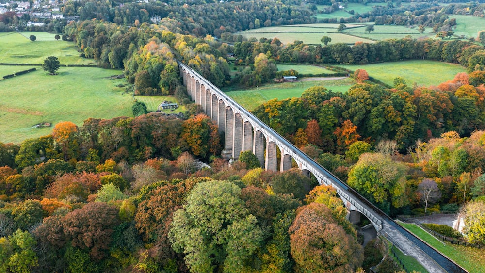 Eine Brücke führt über einen Fluss, umgeben von Herbstlaub und ländlicher Landschaft.