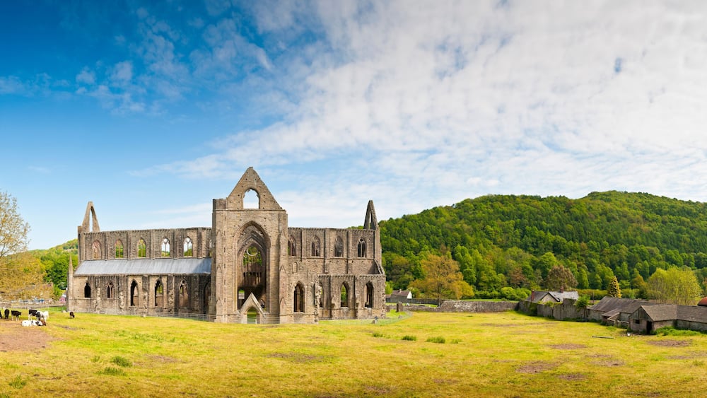 An ancient stone church with a large arched entrance and multiple smaller arches, surrounded by a grassy field and a forested hill in the background.