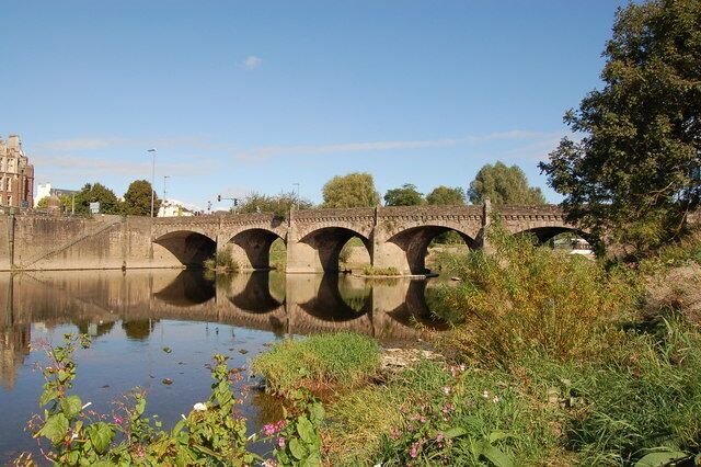 A stone bridge over a calm river with greenery and a building in the background.