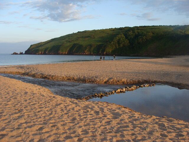 A sandy beach with a rocky water channel, a grassy hill in the background, and a few people in the distance.