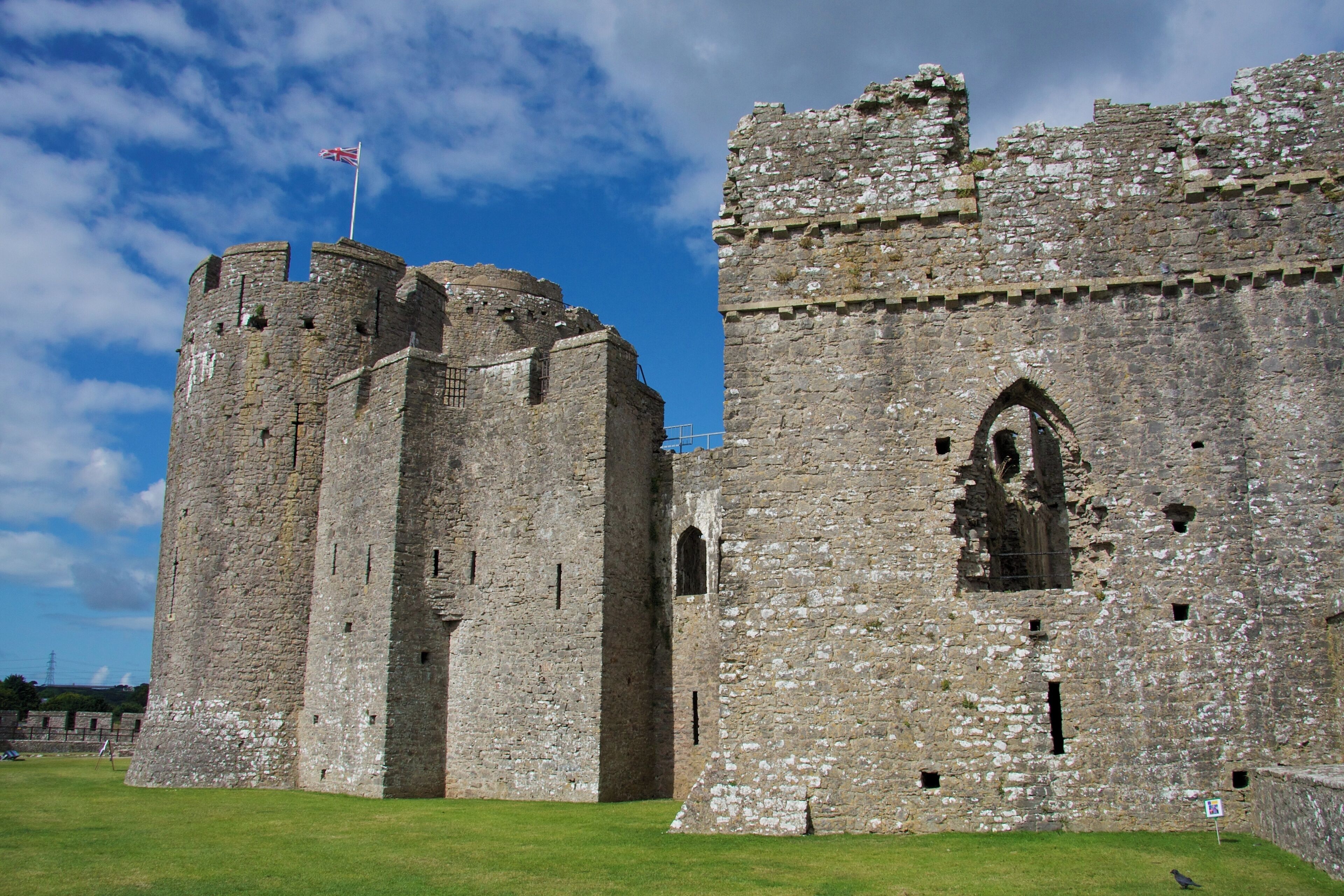 A historic stone castle with a flag on top, a large arched window, and a grassy foreground.
