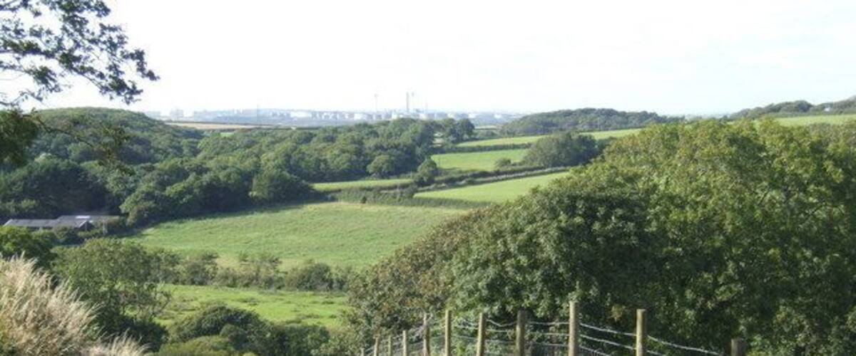 A rural dirt path leading through a green field with a fence on one side and a hilly landscape in the distance.