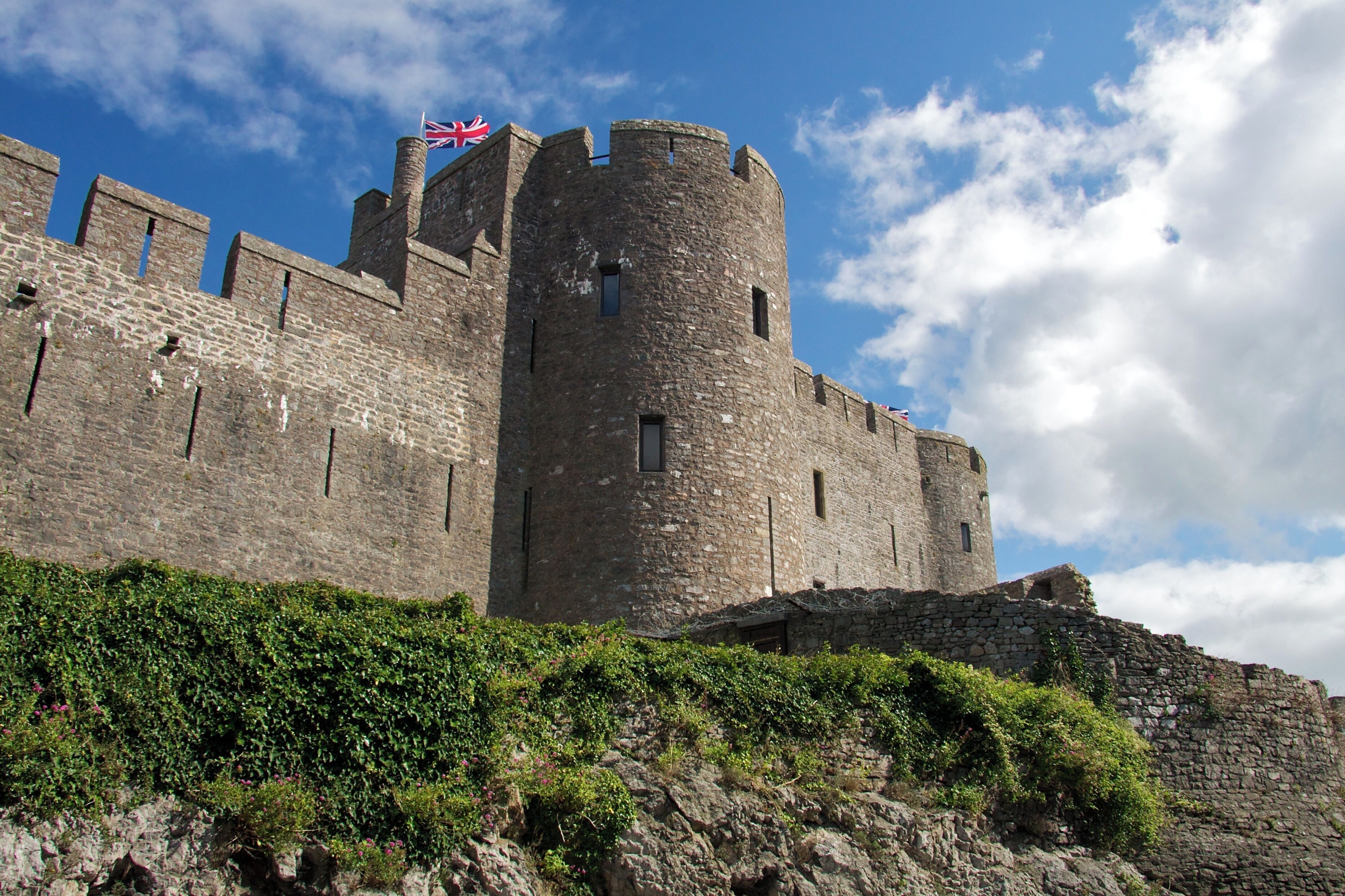 A historic stone castle with a flag, surrounded by greenery and under a partly cloudy sky.