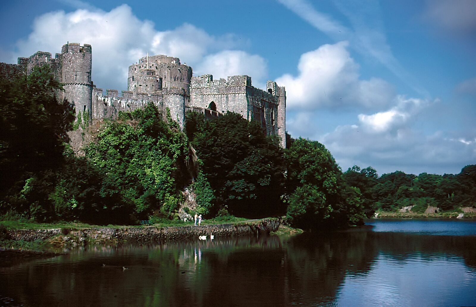 Um castelo histórico numa colina com vista para um lago calmo, rodeado por vegetação exuberante.